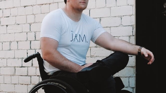 An image of Jason - a white 25 year old male - sitting in his wheelchair. He has short messy brown hair, and is wearing black sunglasses, black boots, black jeans and a white t-shirt with the word JAM on it. He has a calm expression on his face.