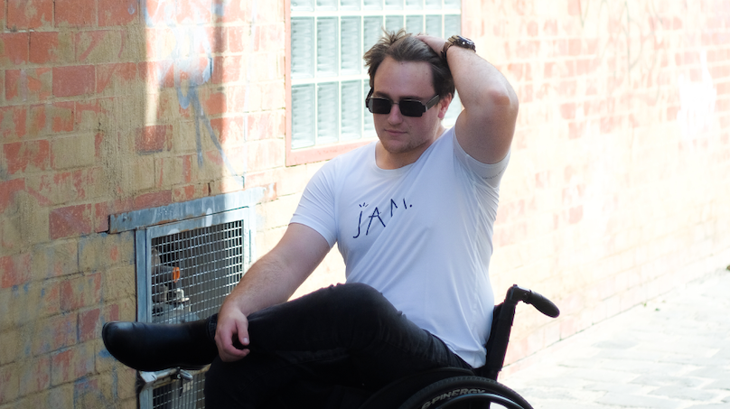 An image of Jason, a man in his 20's wearing sunglasses, a white tshirt with the word JAM written on it, black jeans and black boots, sitting in his wheelchair. He is positioned with a brick wall in the background and is pushing his brown hair back.