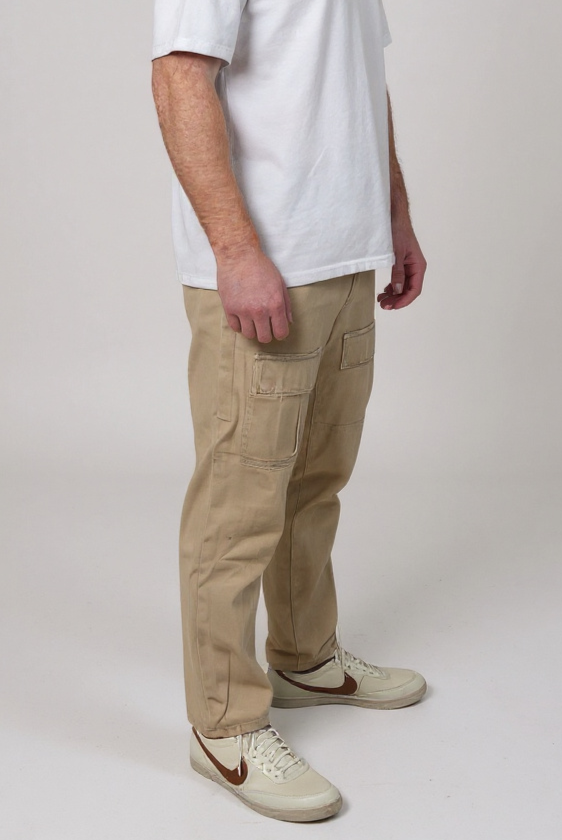 A man visible from the chest down wearing a white t-shirt and beige pants standing against a plain background
