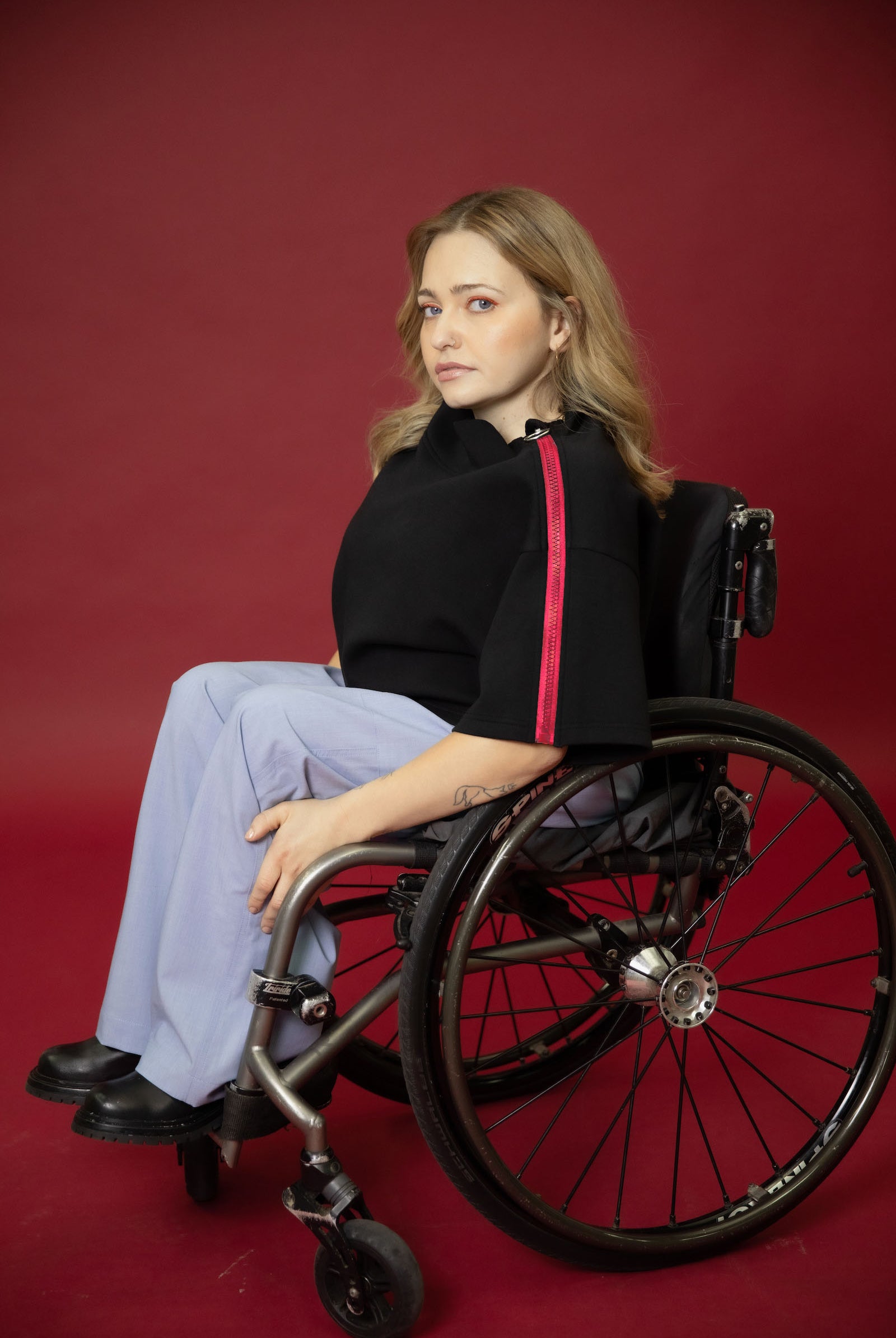 A model seated in her wheelchair side on in front of a maroon background wearing a black cropped top with light blue wide leg pants and black shoes #Seated