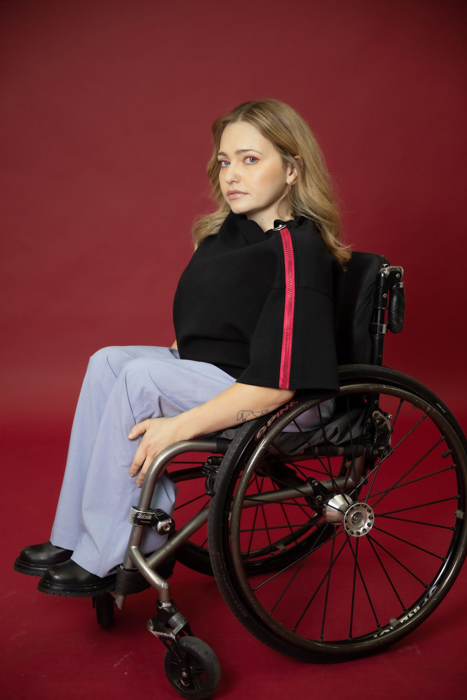 A model seated in her wheelchair side on in front of a maroon background wearing a black cropped top with light blue wide leg pants and black shoes #Seated