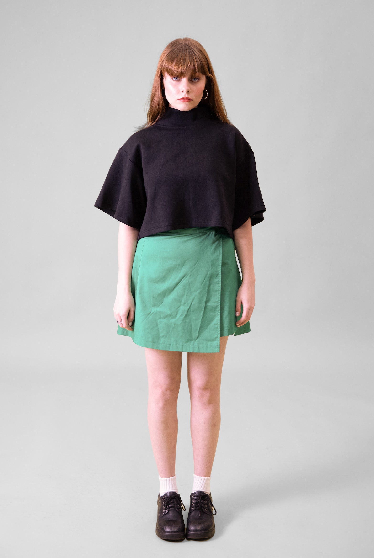 A model standing in front of a grey background wearing a black cropped top and green wrap skirt with black shoes and white socks #Standing