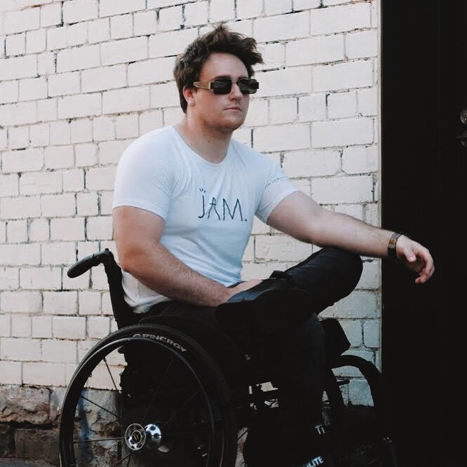 An image of Jason - a white 25 year old male - sitting in his wheelchair. He has short messy brown hair, and is wearing black sunglasses, black boots, black jeans and a white t-shirt with the word JAM on it. He has a calm expression on his face.