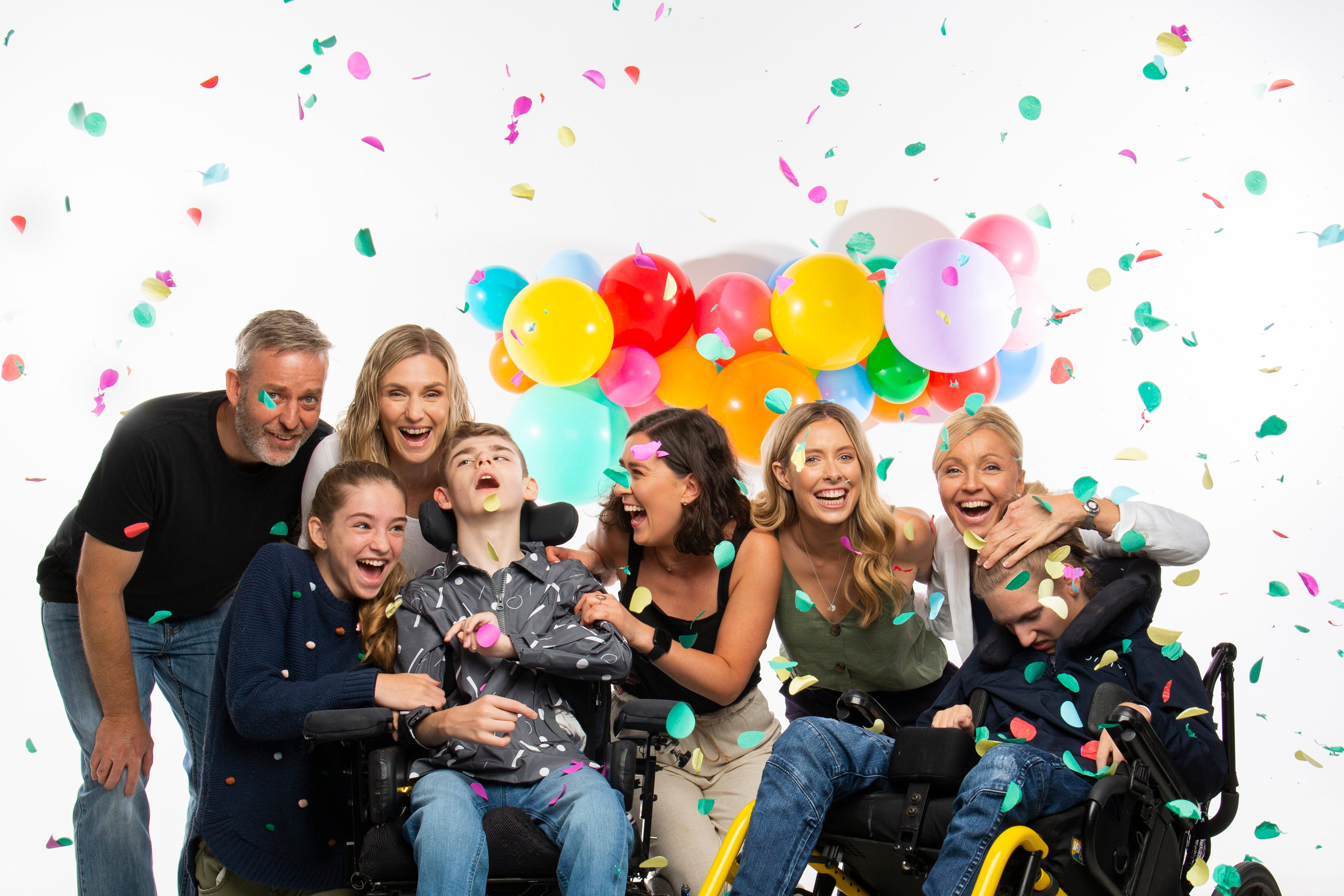 A group of people are surrounded by confetti and there is a brightly coloured balloon garland in the background