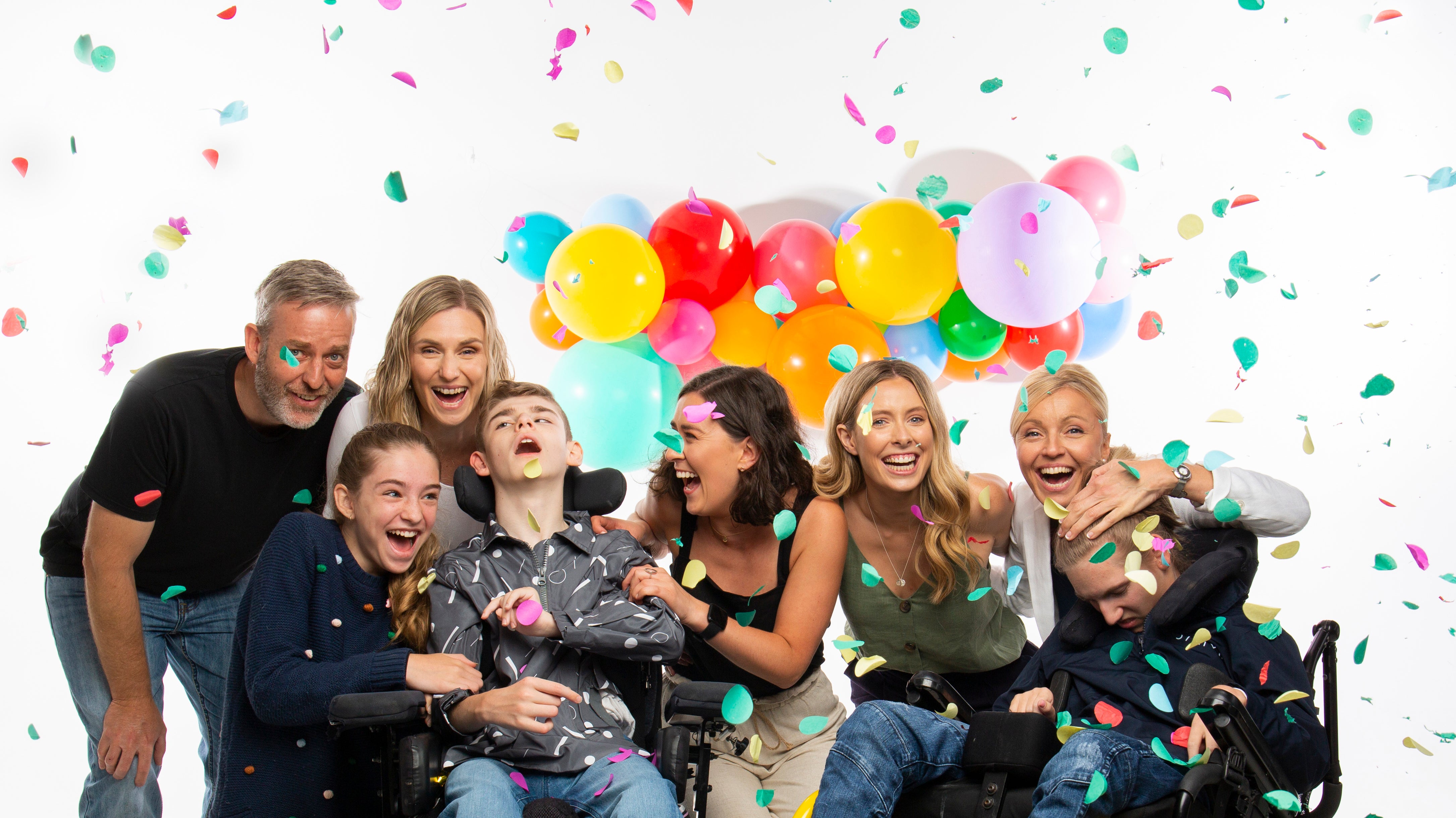 A group of people are surrounded by confetti and there is a brightly coloured balloon garland in the background