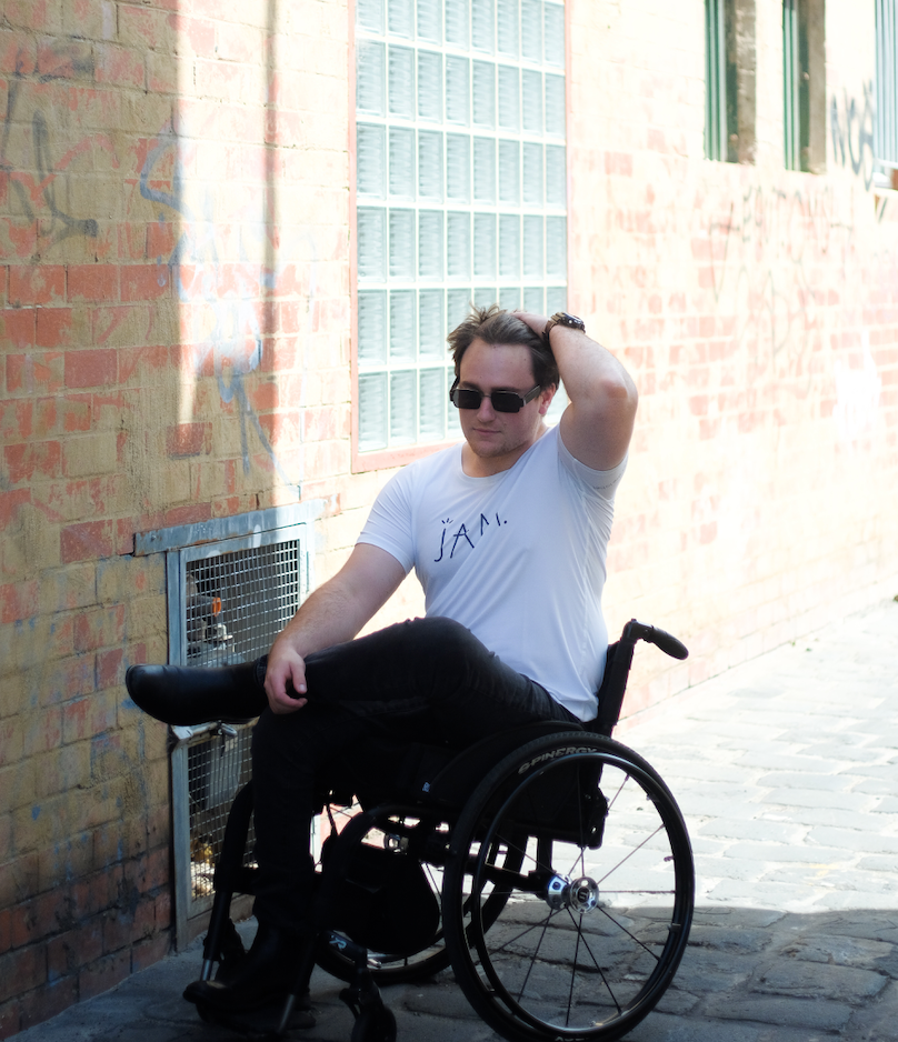 An image of Jason, a man in his 20's wearing sunglasses, a white tshirt with the word JAM written on it, black jeans and black boots, sitting in his wheelchair. He is positioned with a brick wall in the background and is pushing his brown hair back.