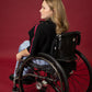A model seated in her wheelchair facing away from the camera in front of a maroon background wearing a black cropped top with light blue wide leg pants and black shoes #Seated