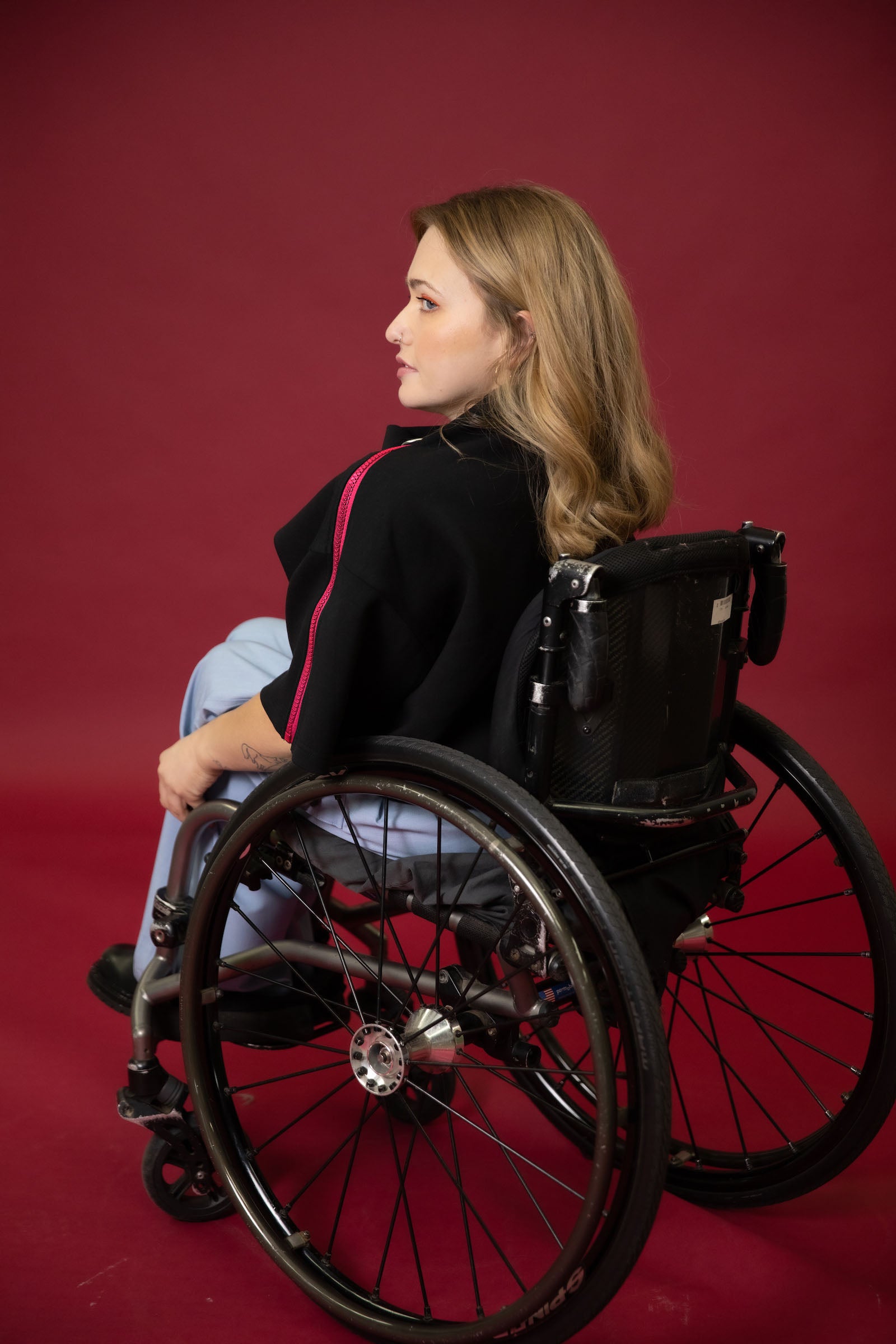 A model seated in her wheelchair facing away from the camera in front of a maroon background wearing a black cropped top with light blue wide leg pants and black shoes #Seated