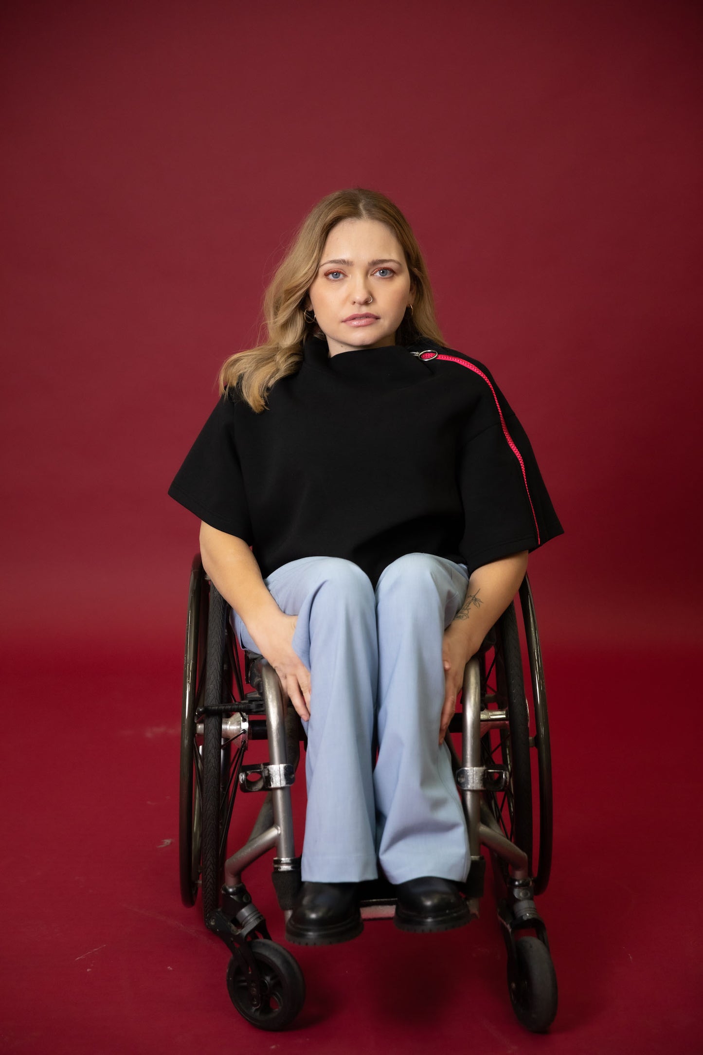 A model seated in her wheelchair in front of a maroon background wearing a black cropped top with light blue wide leg pants and black shoes #Seated