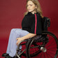 A model seated in her wheelchair side on in front of a maroon background wearing a black cropped top with light blue wide leg pants and black shoes #Seated