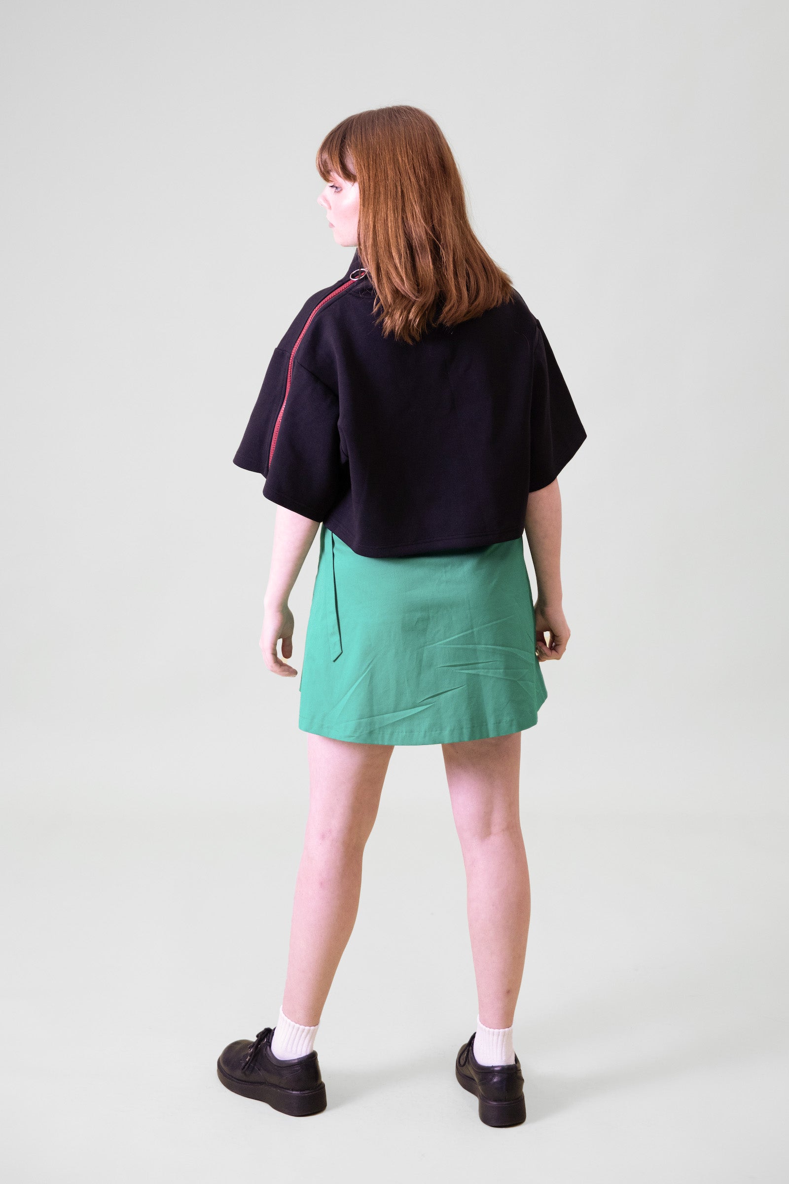 A model standing in front of a grey background facing backwards towards the wall wearing a black cropped top and green wrap skirt with black shoes and white socks #Standing
