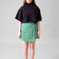A model standing in front of a grey background wearing a black cropped top and green wrap skirt with black shoes and white socks #Standing