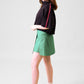 A model standing sideways in front of a grey background facing backwards towards the wall wearing a black cropped top and green wrap skirt with black shoes and white socks #Standing
