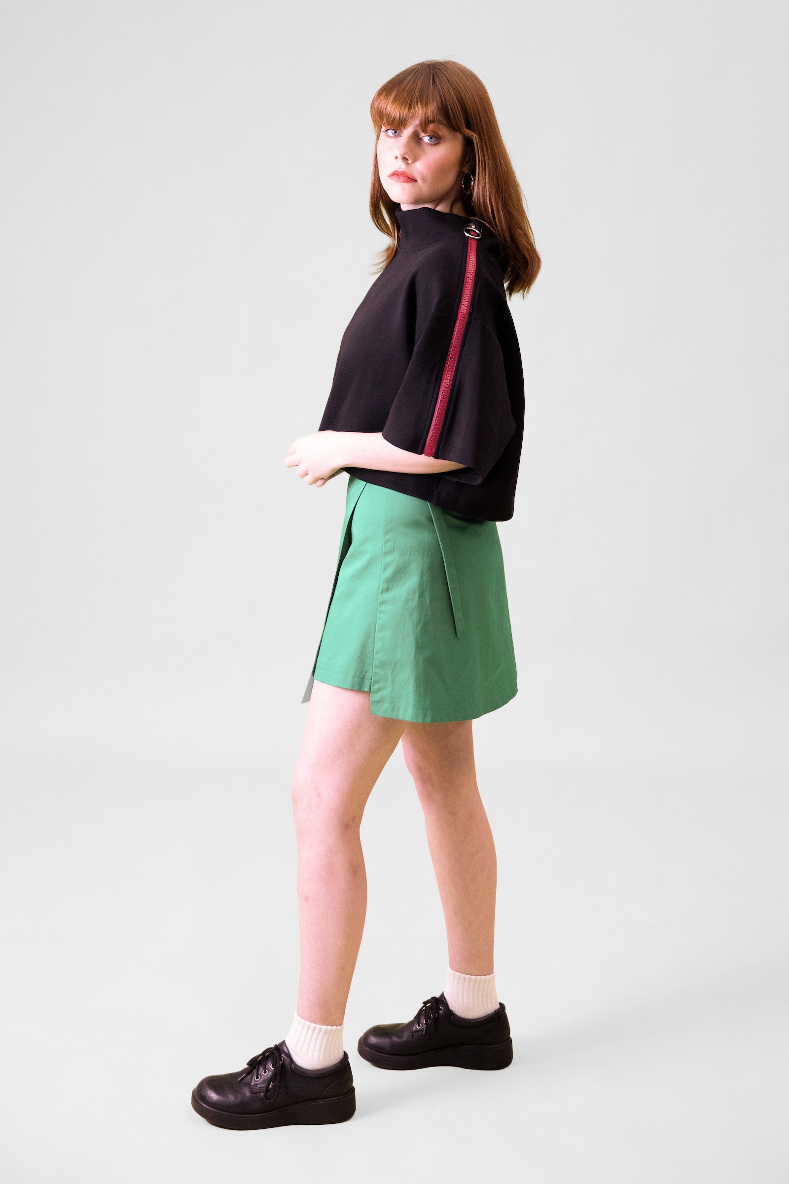 A model standing sideways in front of a grey background facing backwards towards the wall wearing a black cropped top and green wrap skirt with black shoes and white socks #Standing