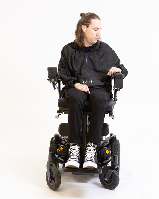 A model seated in his wheelchair in front of a white background. He is wearing a black jacket with black chinos and sneakers. #Seated