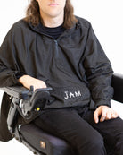 A close up image of a model seated in his wheelchair in front of a white background. He is wearing a black windbreaker jacket with black chinos. #Seated