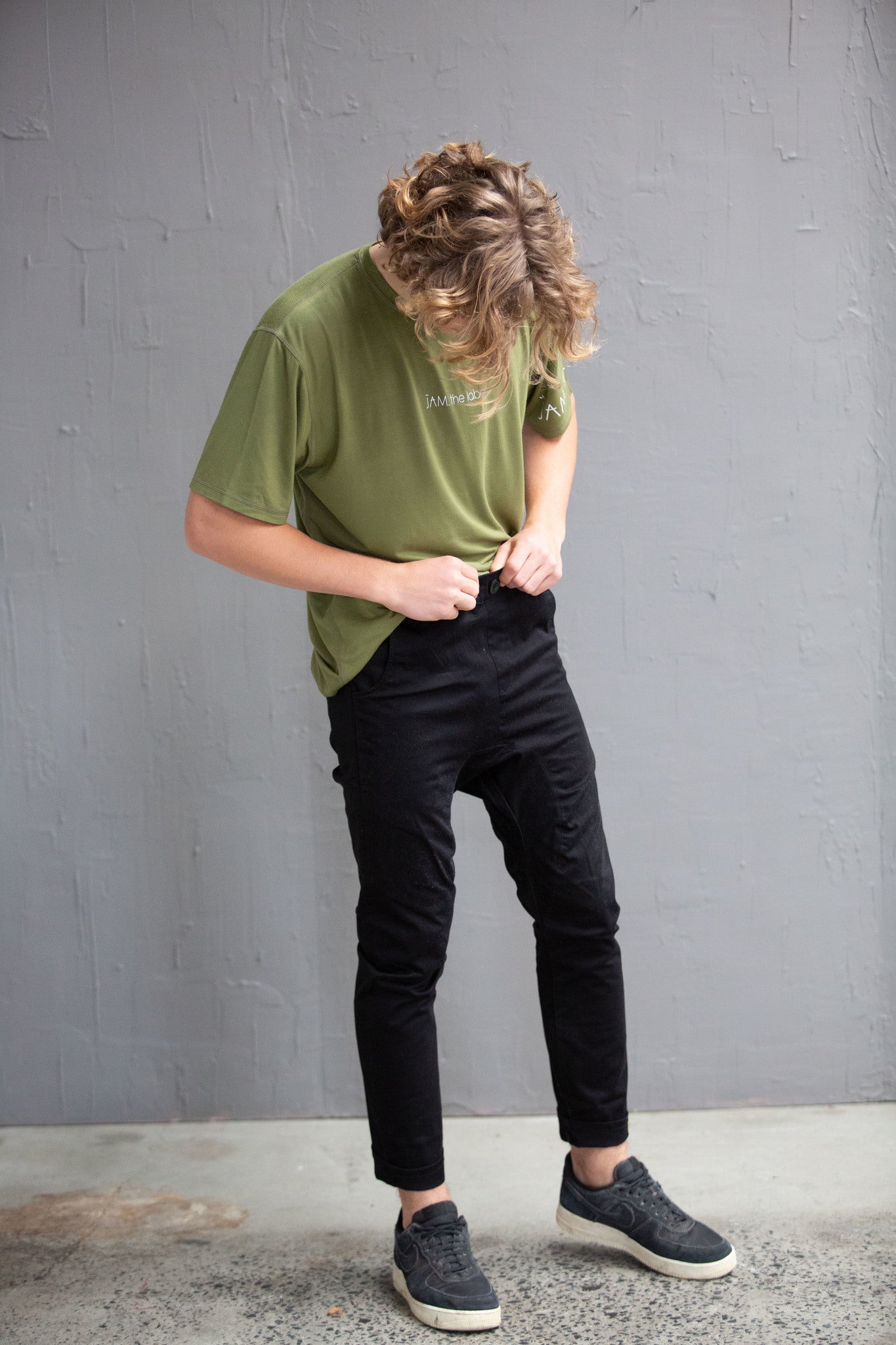 A young white man with blonde curly hair is standing in front of a grey wall. He is looking down at himself. He is wearing an olive green top, black chinos and black shoes.