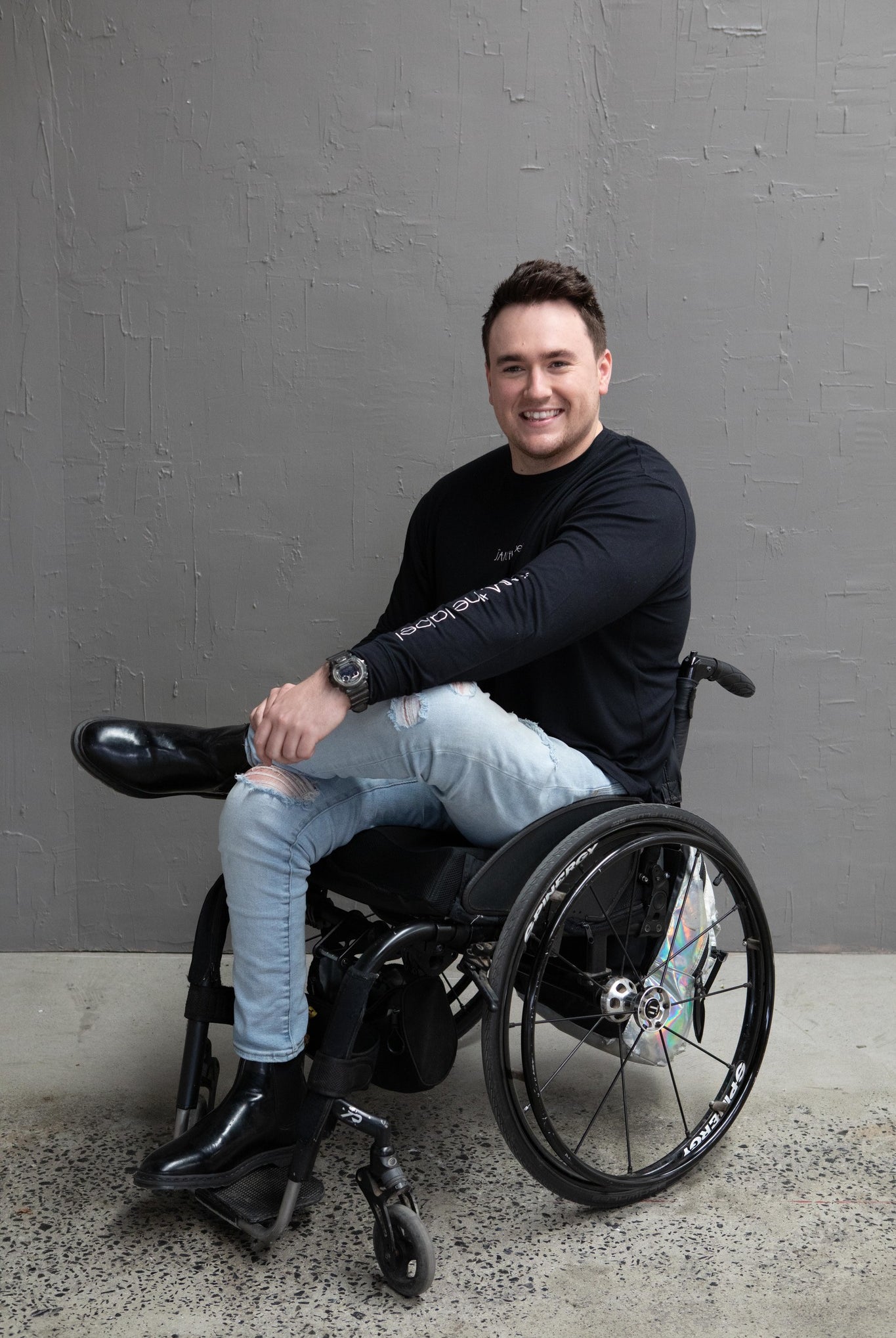 Jason, a white man with short brown hair, is sitting in his wheelchair and smiling. He is wearing a black long sleeve with the words "JAM the label" down the sleeve, ripped light blue jeans and black boots. One leg is crossed over the other. He is against a grey wall and the floor is concrete.