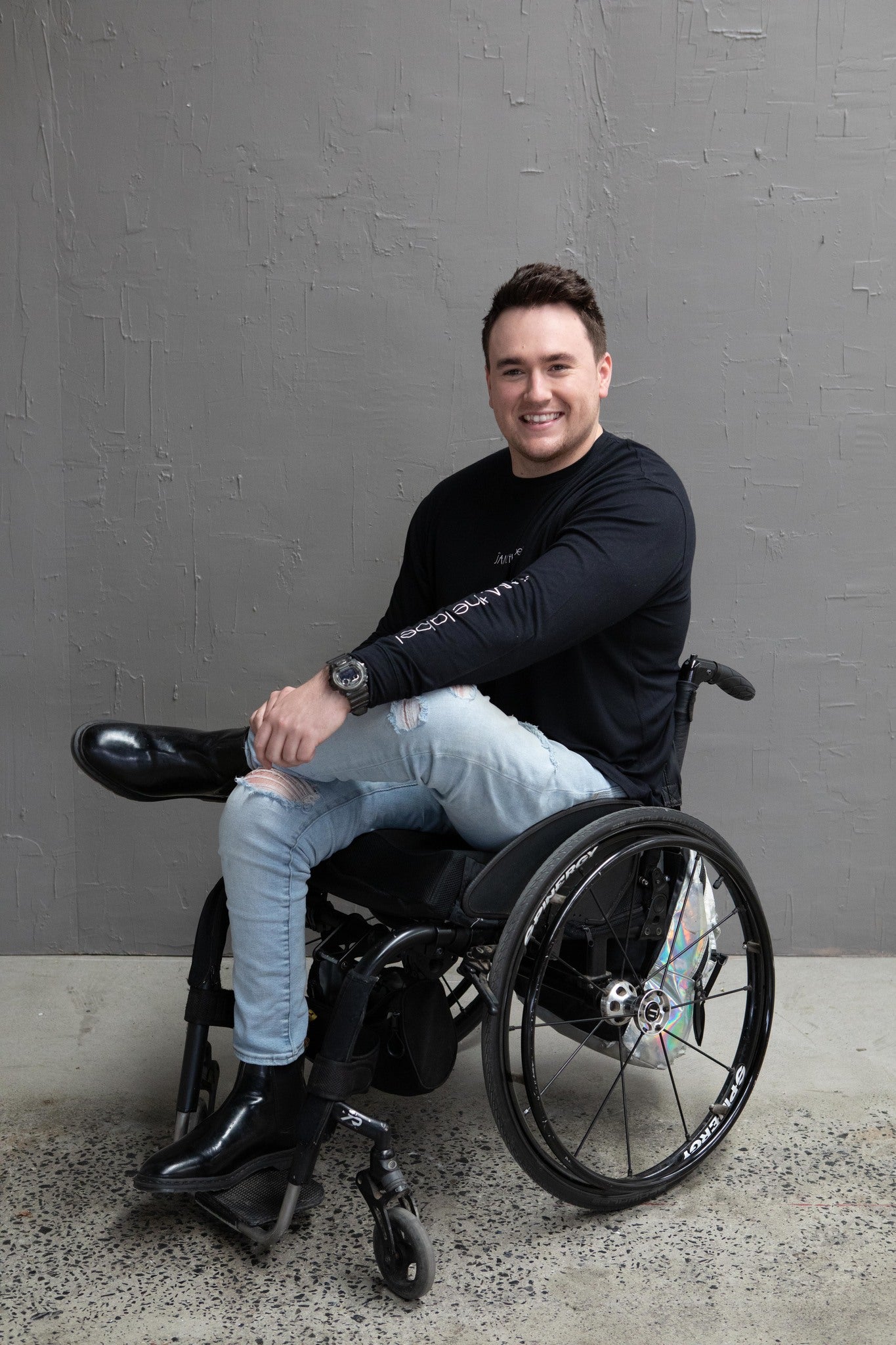 Jason, a white man with short brown hair, is sitting in his wheelchair and smiling. He is wearing a black long sleeve with the words "JAM the label" down the sleeve, ripped light blue jeans and black boots. One leg is crossed over the other. He is against a grey wall and the floor is concrete.
